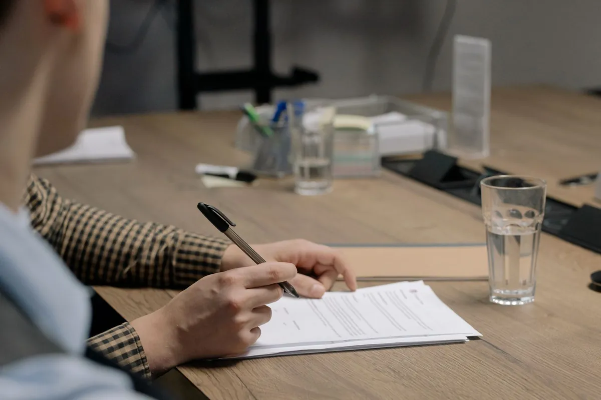 A professional in business attire signs a document at a wooden table in an office setting.
