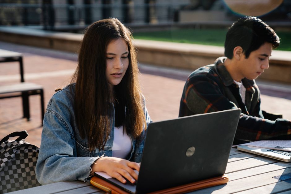 Two young adults working on laptops at an outdoor table.